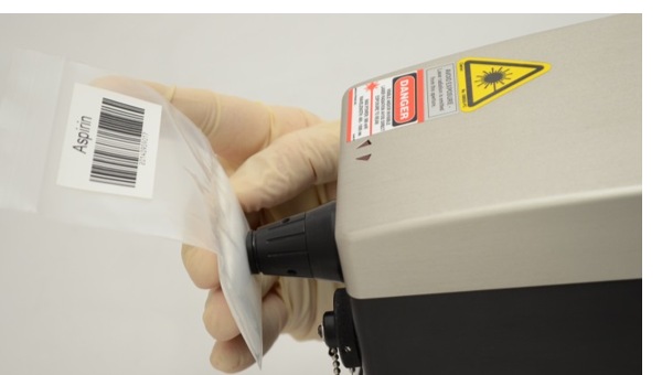 Main gantée pressant un sachet scellé étiqueté contre le nez d’un spectromètre RAMAN portable ; l’étiquette de sécurité laser est visible sur le boîtier.